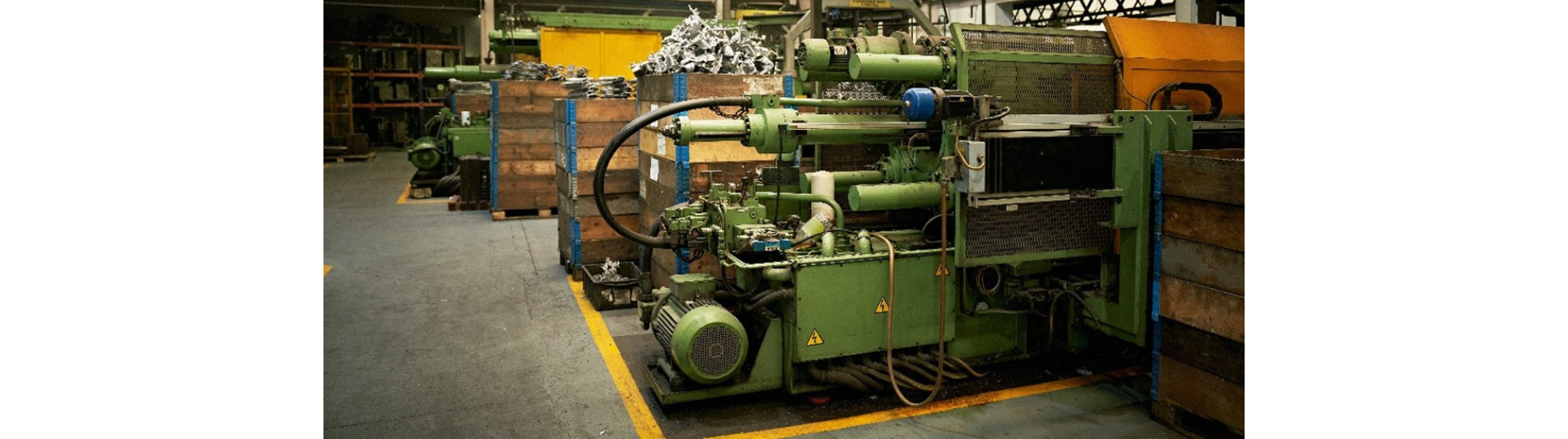 A row of large green industrial machines sits inside a manufacturing facility, surrounded by wooden crates and metal bins. Yellow floor markings outline walkways, and hydraulic hoses and control panels are visible on the equipment.